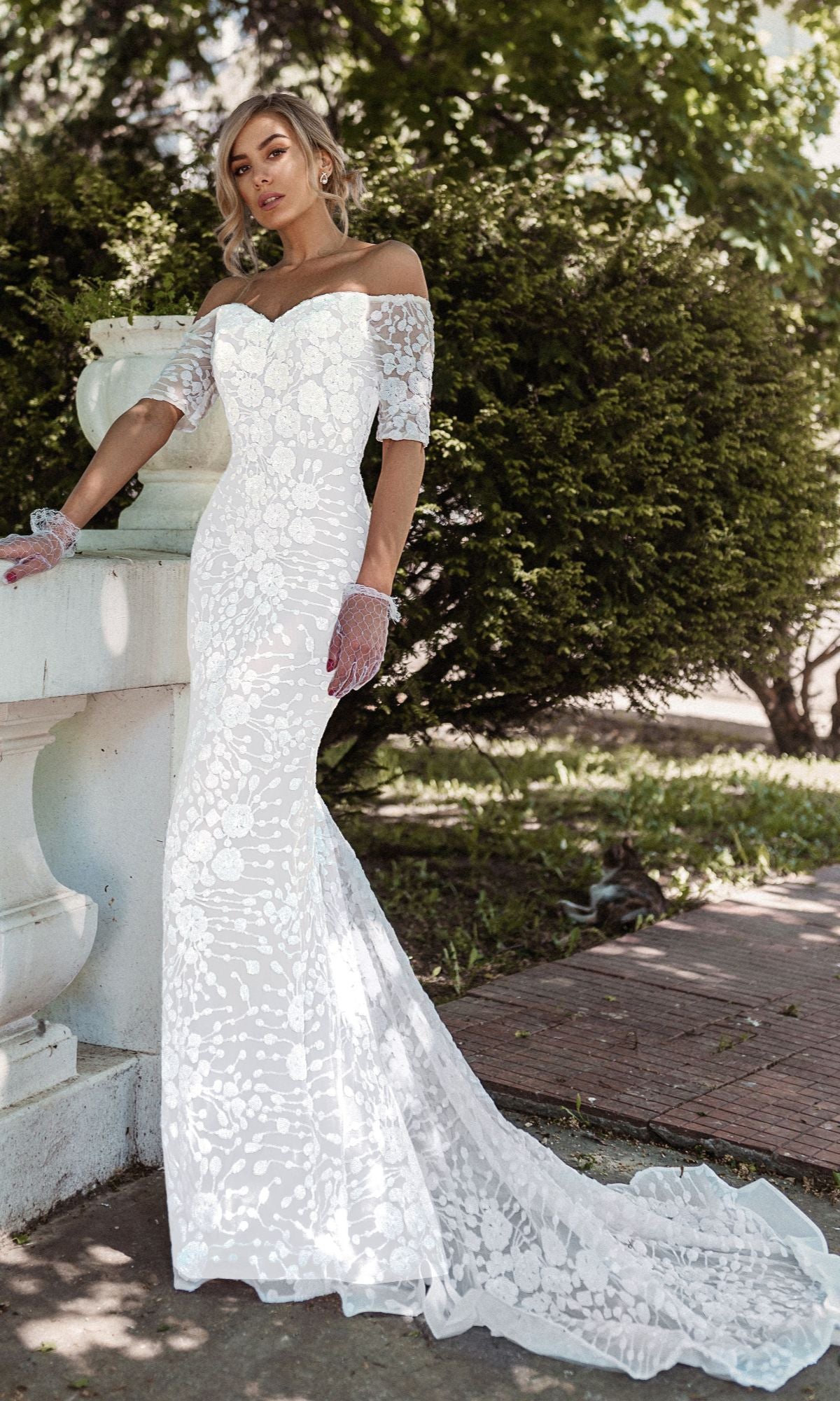 A woman stands outdoors in the Off-Shoulder White Bridal Gown: Tina Holly BB009—a fitted lace gown with a long train and lace gloves—surrounded by greenery and elegant stone railings.